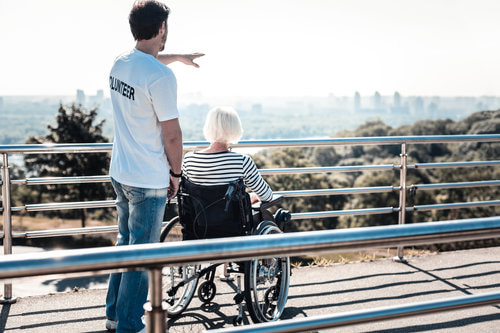 A male standing in back of an old lady who is sitting in a wheelchair and looking at the nice city view.